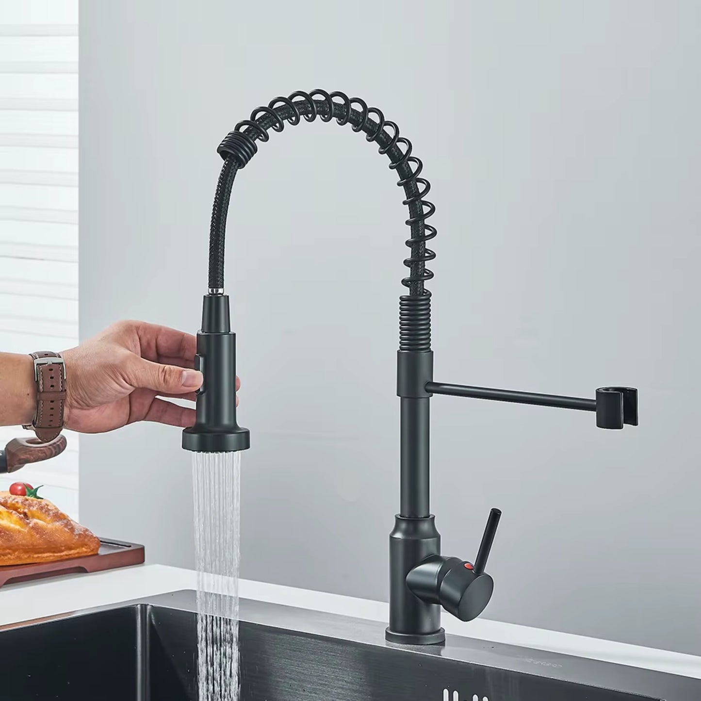 Person holding the pull-down spray head of a matte black kitchen faucet with water flowing into a sink, in a modern setting.